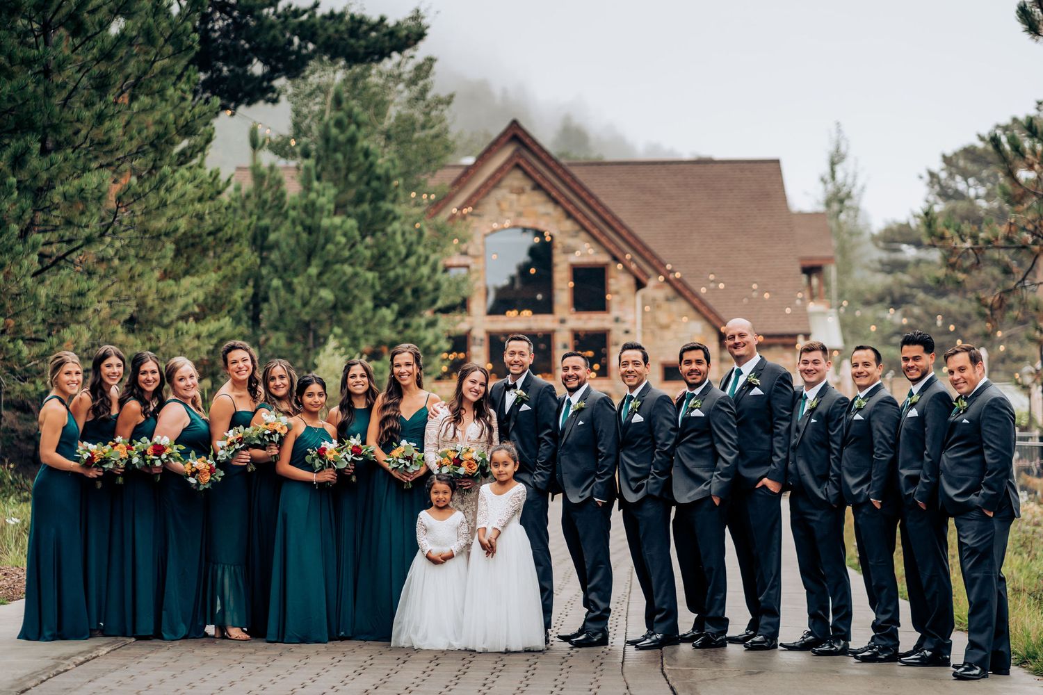 Wedding party in teal dresses and navy suits lines up symmetrically in front of rustic venue.
