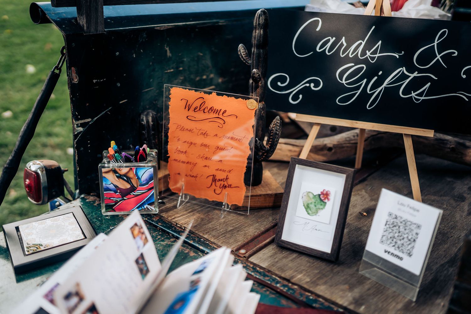 Wedding cards and gifts table with chalkboard sign and framed photos on rustic wooden surface.