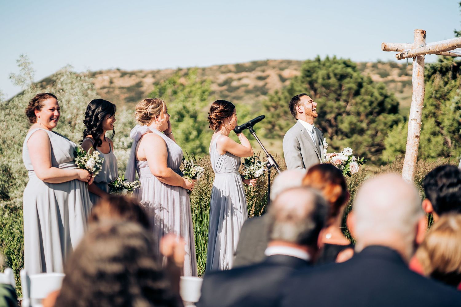 Wedding party standing at rustic wooden altar with mountains and blue sky in background.