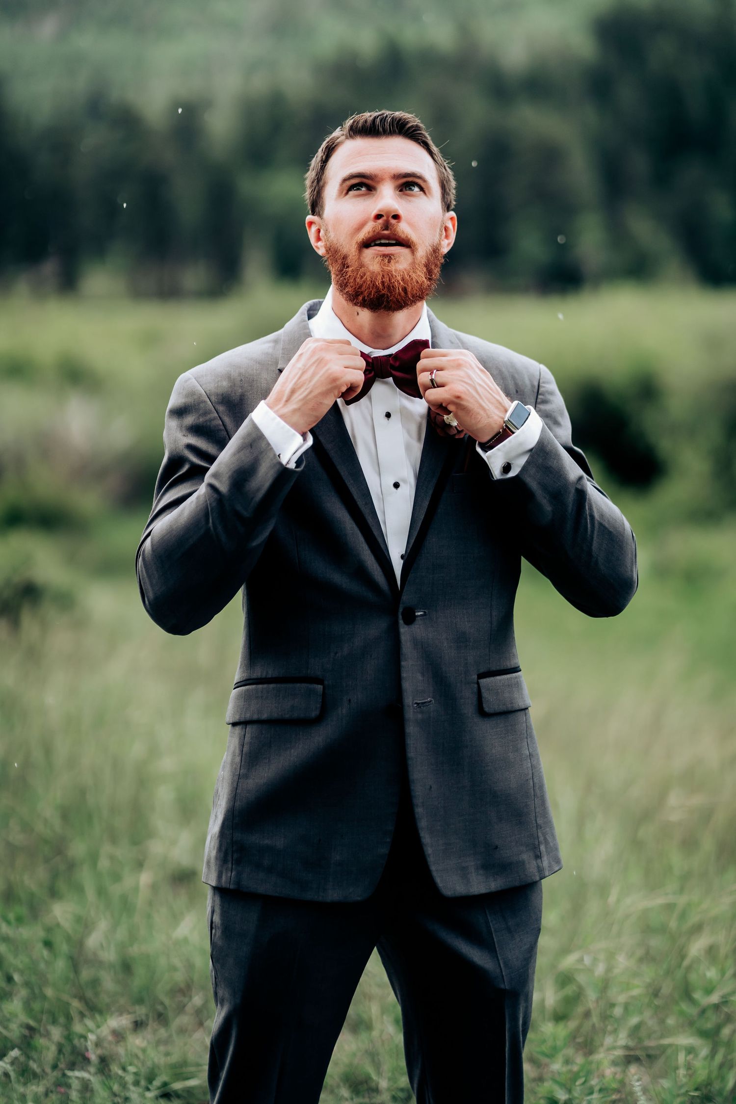 Groom adjusts his bow tie while wearing a classic grey suit outdoors.