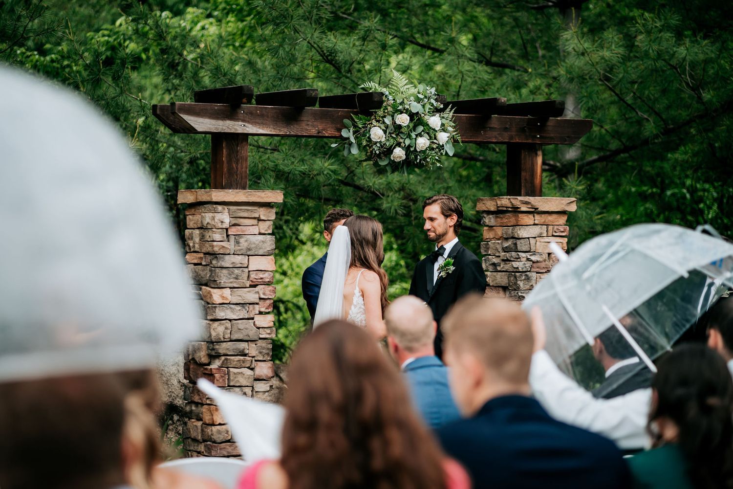 Wedding ceremony under a wooden arch with stone pillars and white floral arrangements.