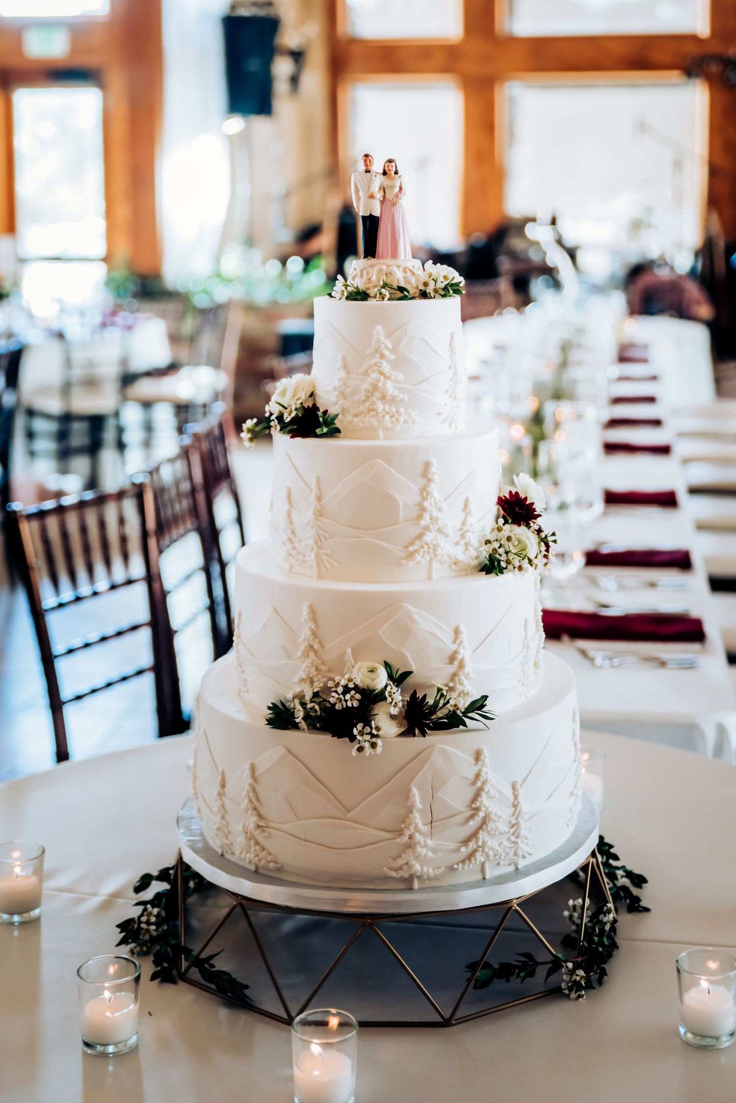 Four-tier white wedding cake decorated with pine trees and floral accents on elegant display.
