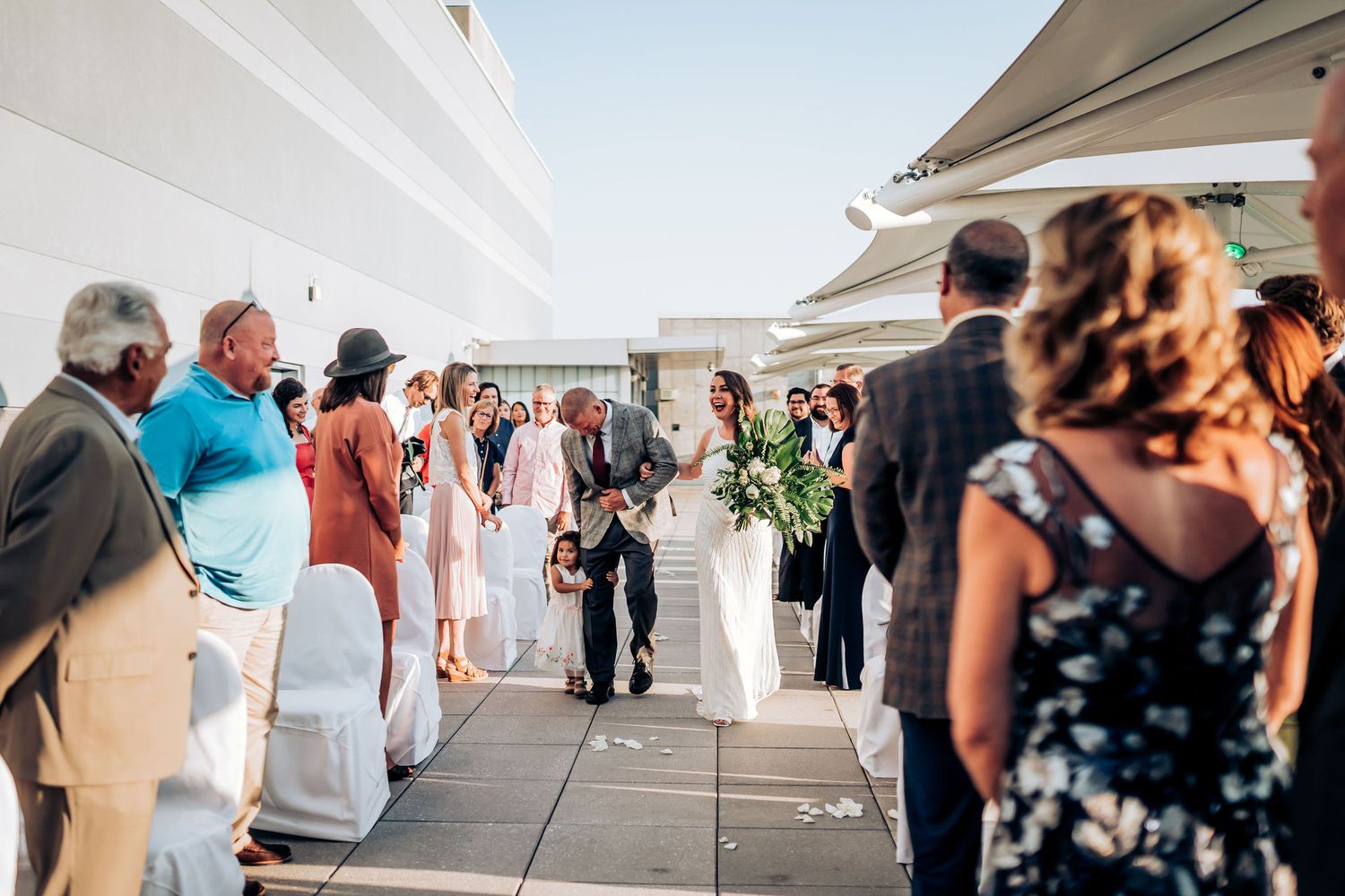 Wedding ceremony processional on a bright rooftop terrace with guests seated in white chairs.