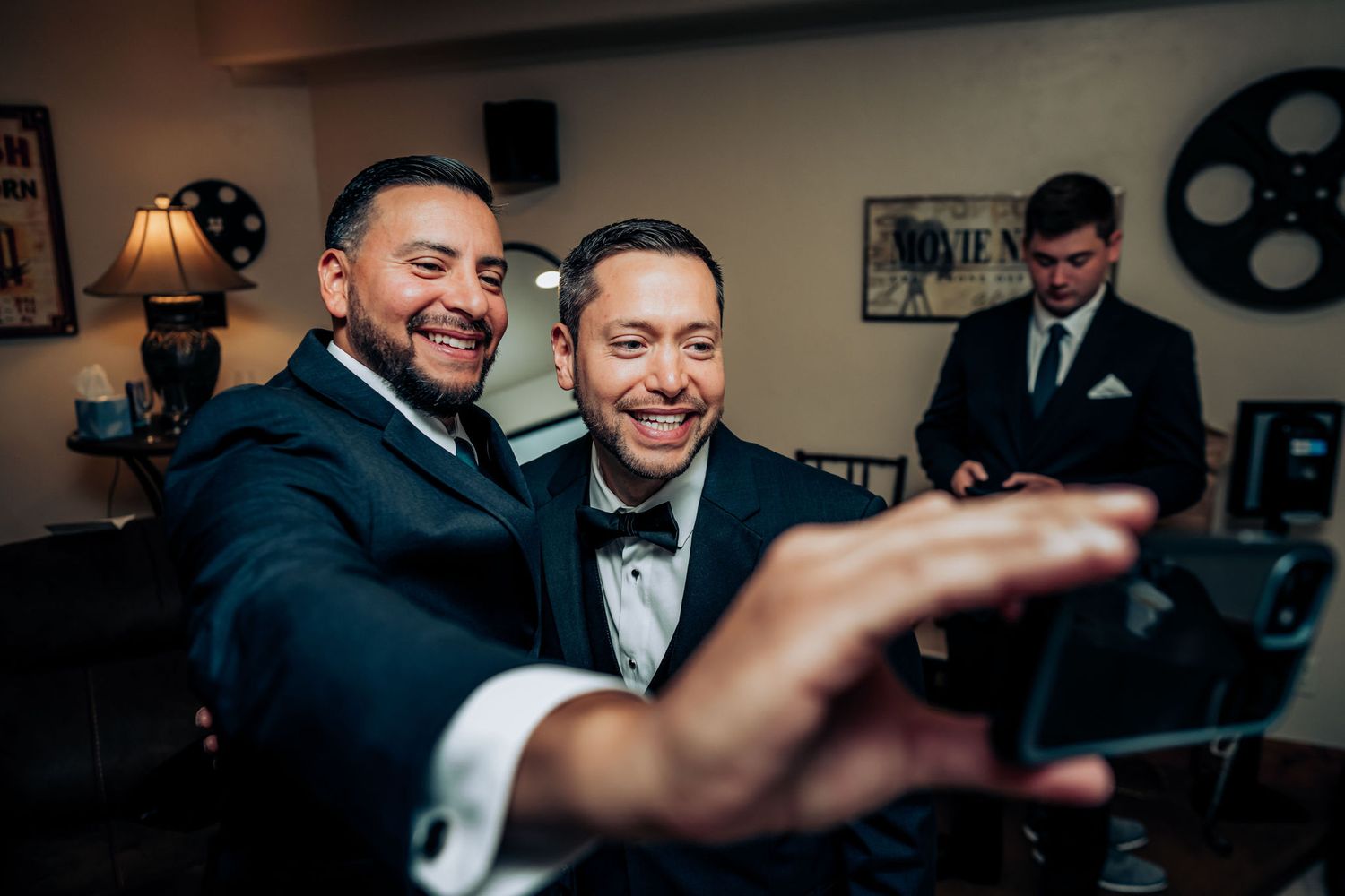 Two groomsmen in black tuxedos share a candid moment before a wedding celebration.