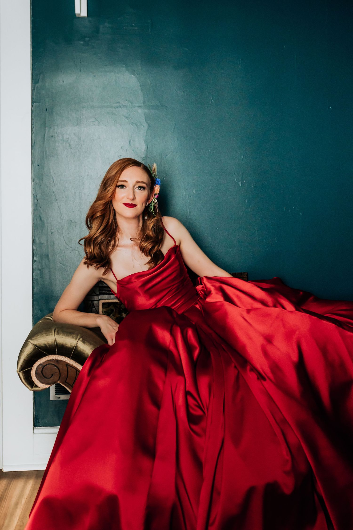 Woman in flowing red satin gown poses dramatically on vintage sofa against teal wall.