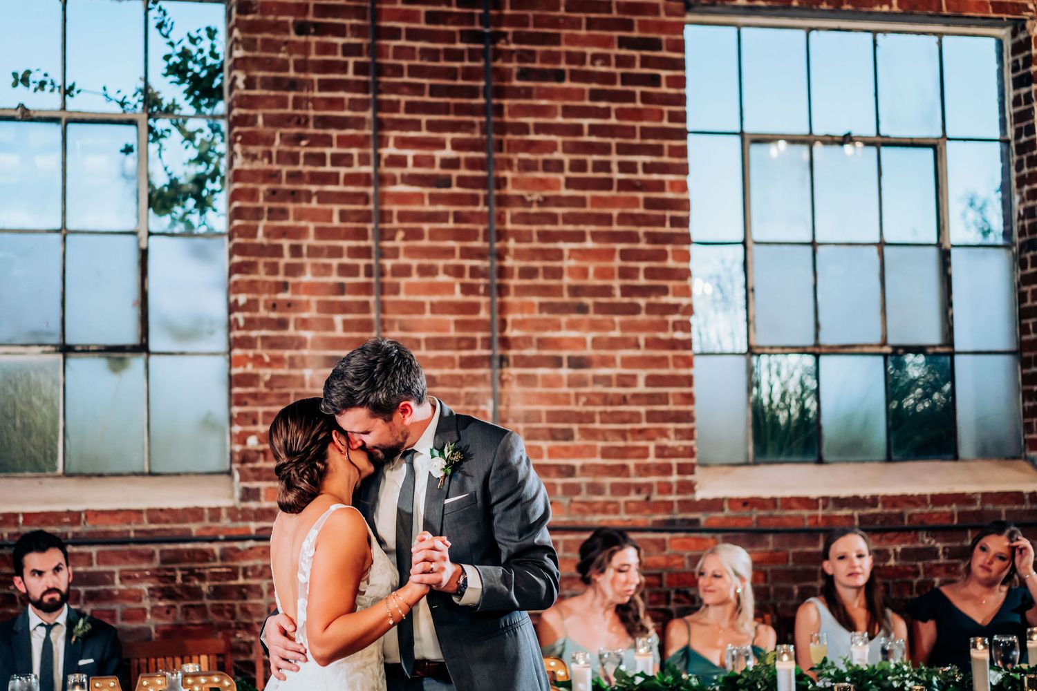 A couple shares their first dance in front of a rustic brick wall with wedding party members seated at a table.