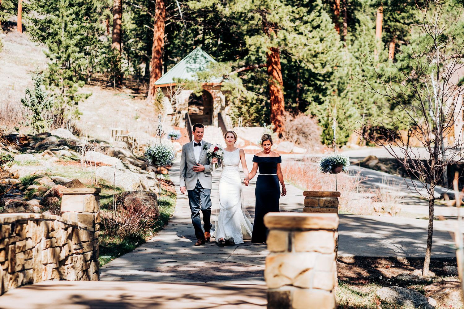 Wedding ceremony procession walking down stone-lined pathway in mountain forest setting.