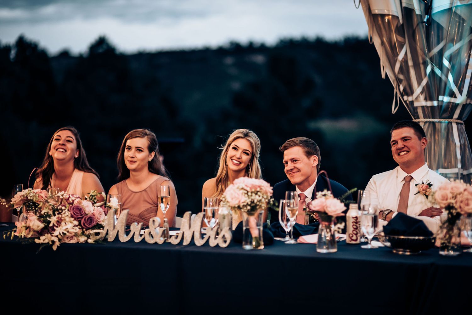 Wedding party members seated at head table laughing during reception speeches.