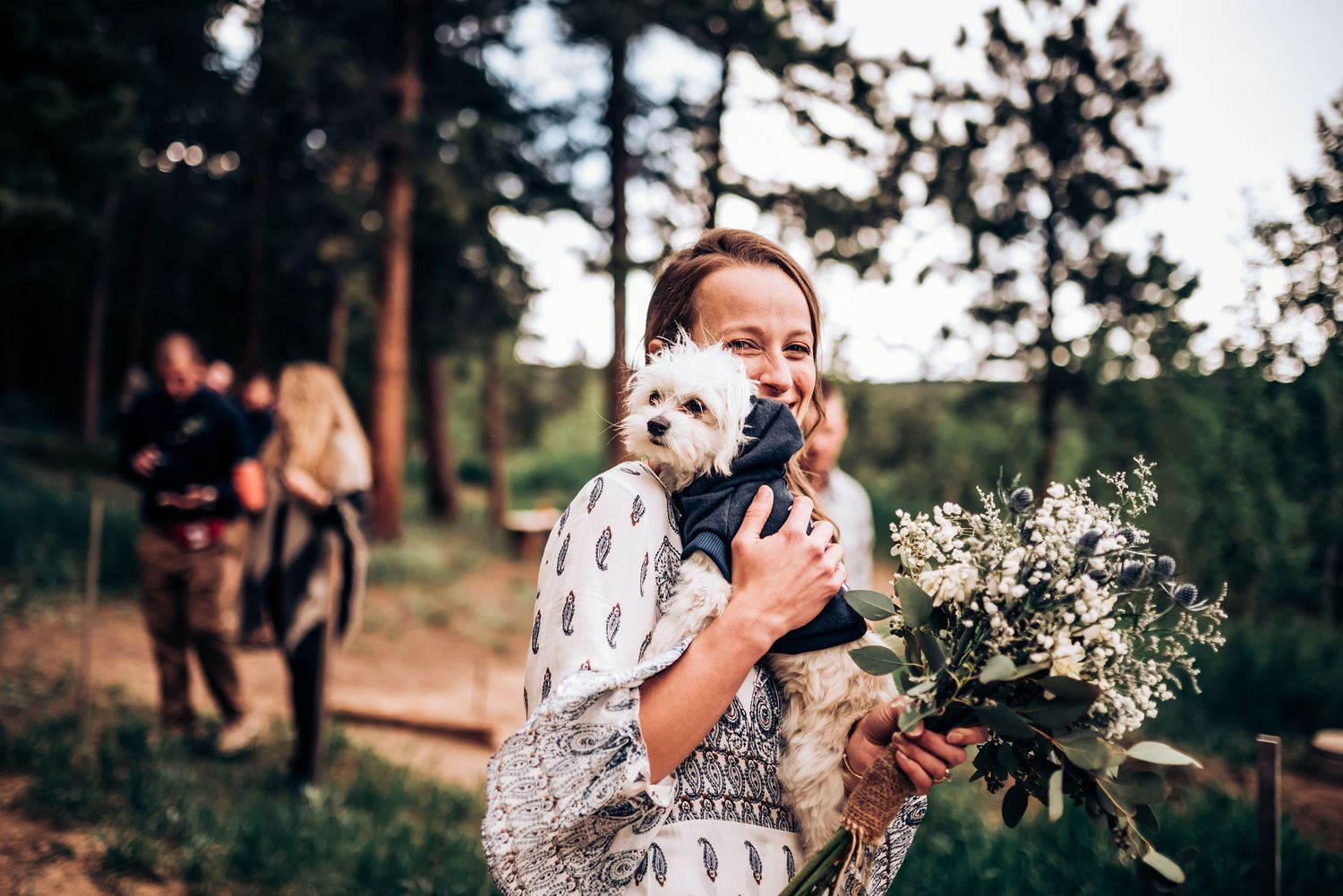 A floral bouquet and small white dog are held outdoors with pine trees in the background at sunset.