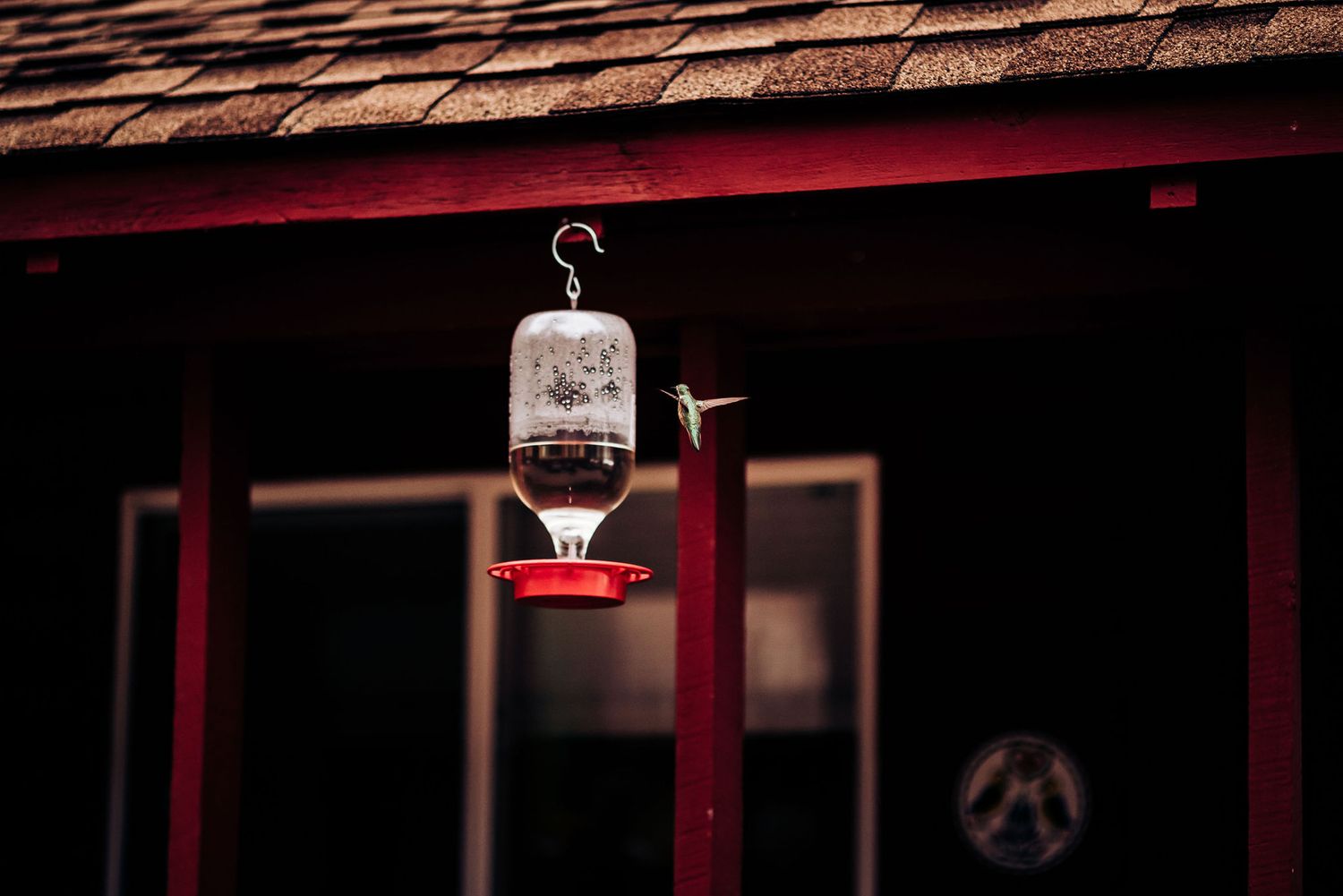 Vintage-style hummingbird feeder hanging from wooden cabin roof at dusk.