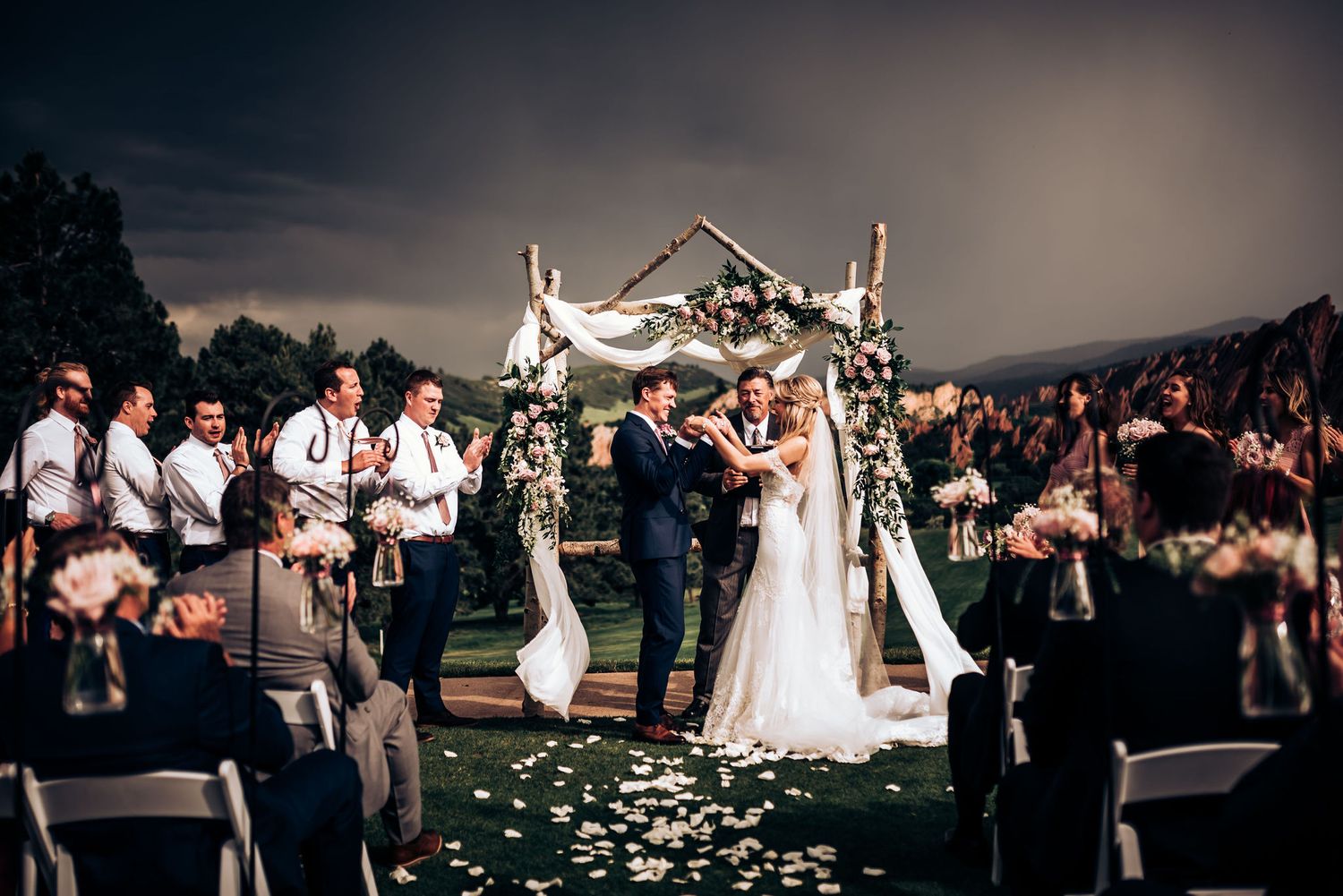 Outdoor wedding celebration with white floral arch and scattered petals on grassy aisle at sunset.
