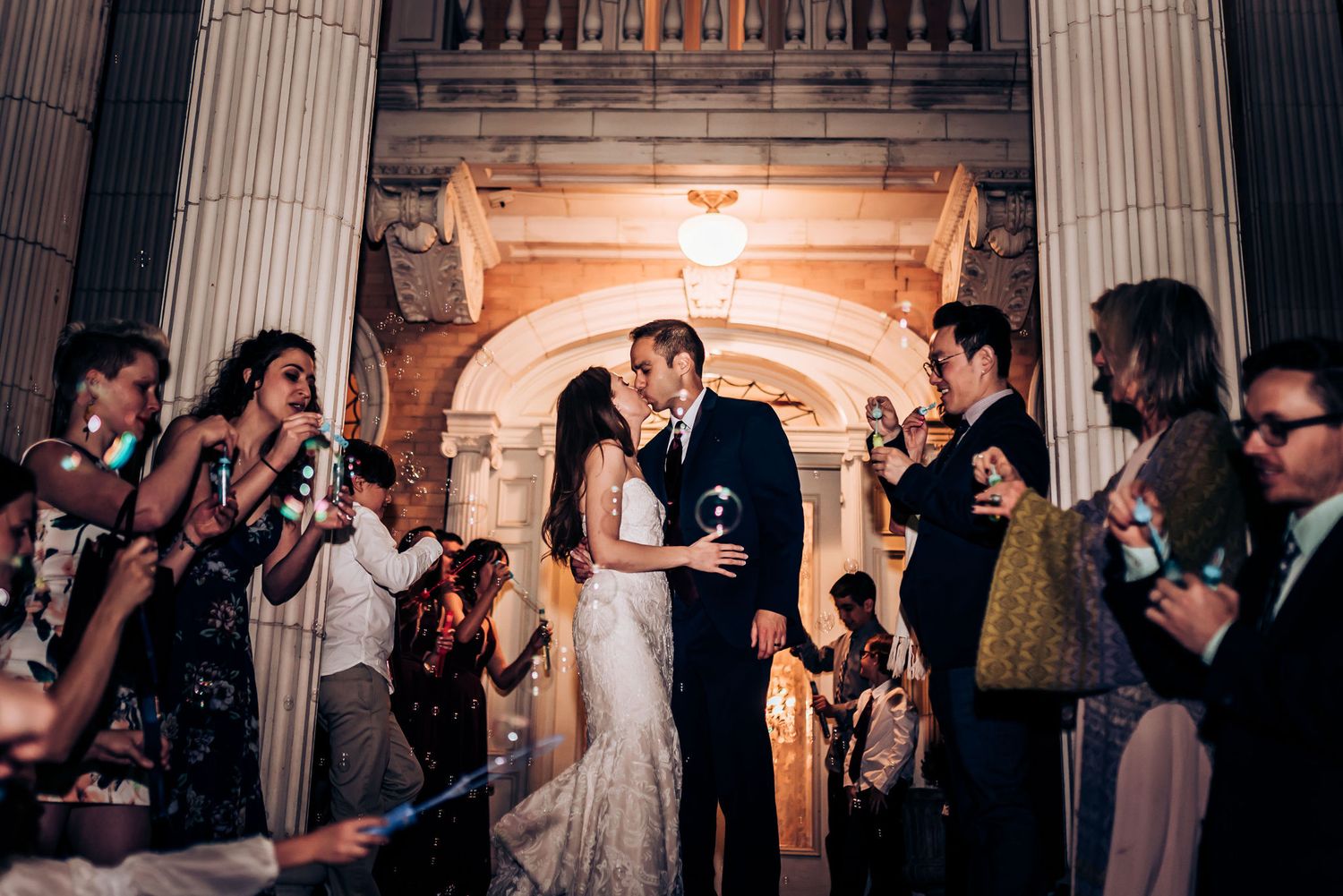 Couple sharing romantic moment under grand archway as guests celebrate with sparklers.