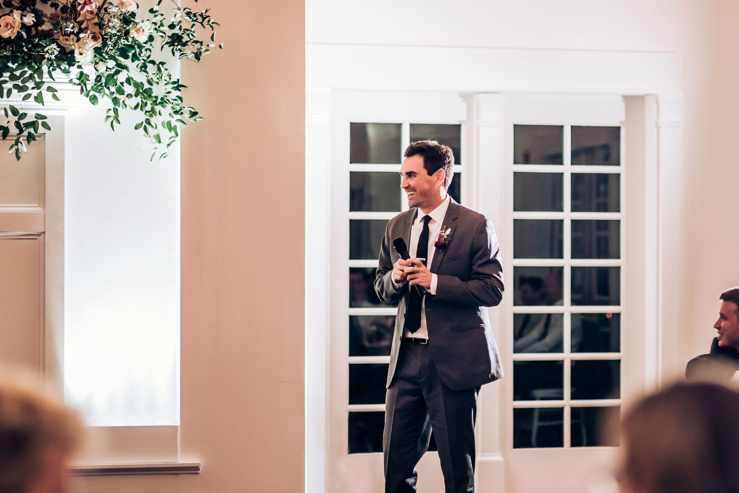 Person in dark suit giving a speech at an elegant indoor wedding reception venue with white walls and greenery decor.