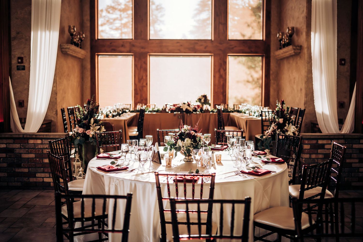 Elegant wedding reception venue with round tables set with white linens and wooden chairs in a rustic indoor setting.