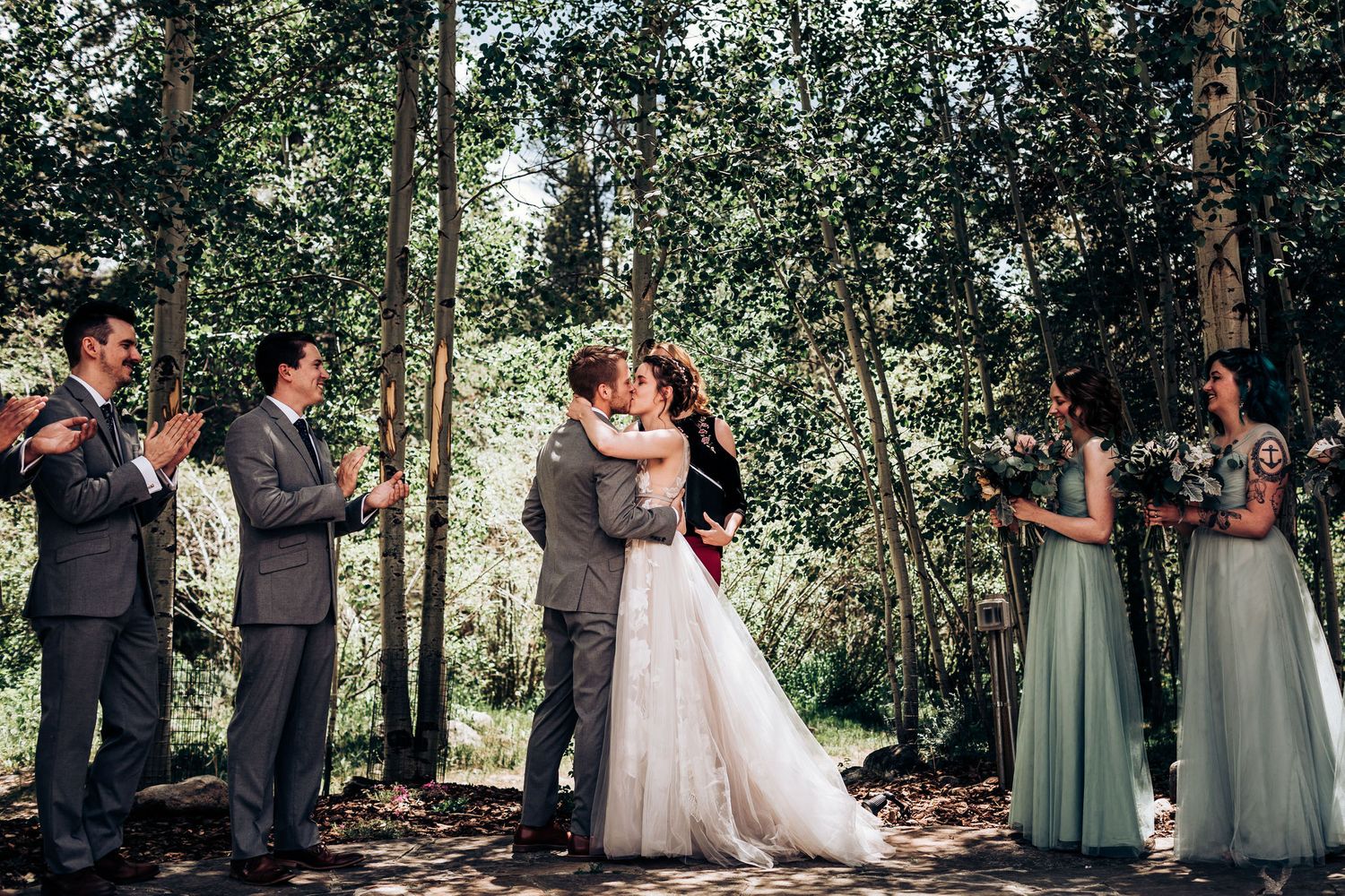 Wedding party applauds as newlyweds share their first kiss in a forest ceremony setting.