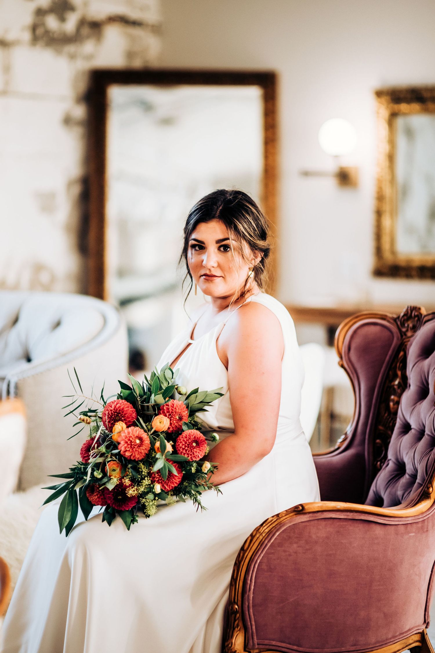 Person in white wedding dress sits in vintage velvet chair holding vibrant coral and green bouquet.