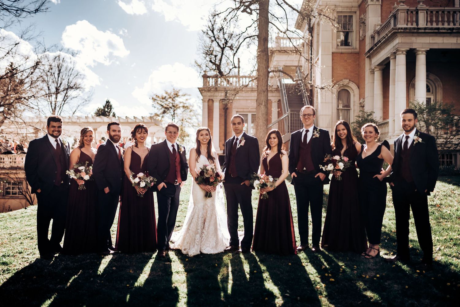Wedding party in dark formal attire poses in silhouette against sunlit historic mansion backdrop.