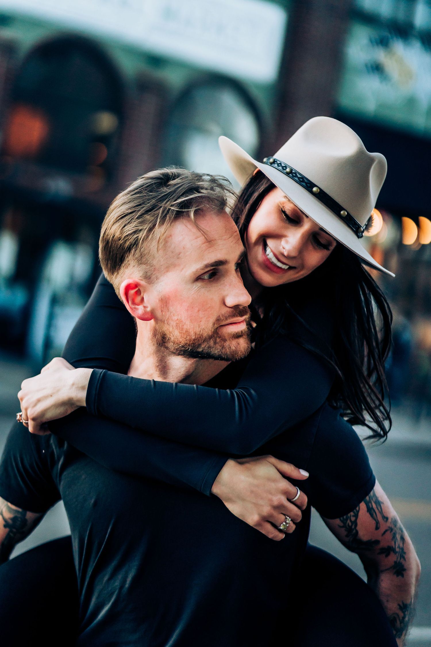 A couple embraces outdoors with one wearing a stylish fedora hat.