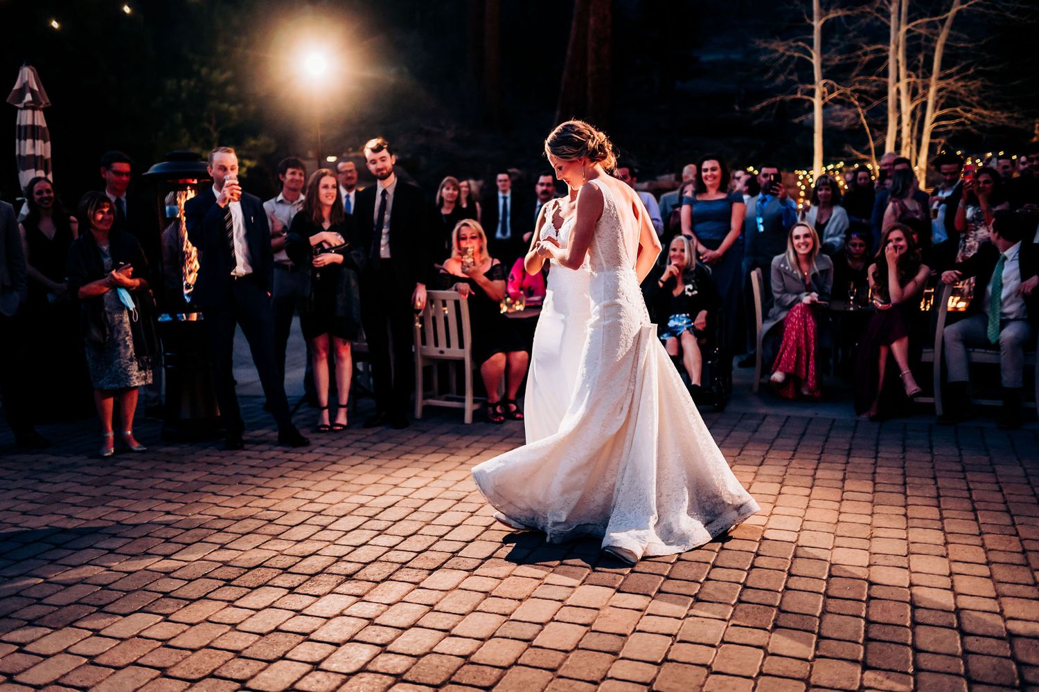Two brides share an intimate dance moment on a brick patio while guests watch from the sidelines.