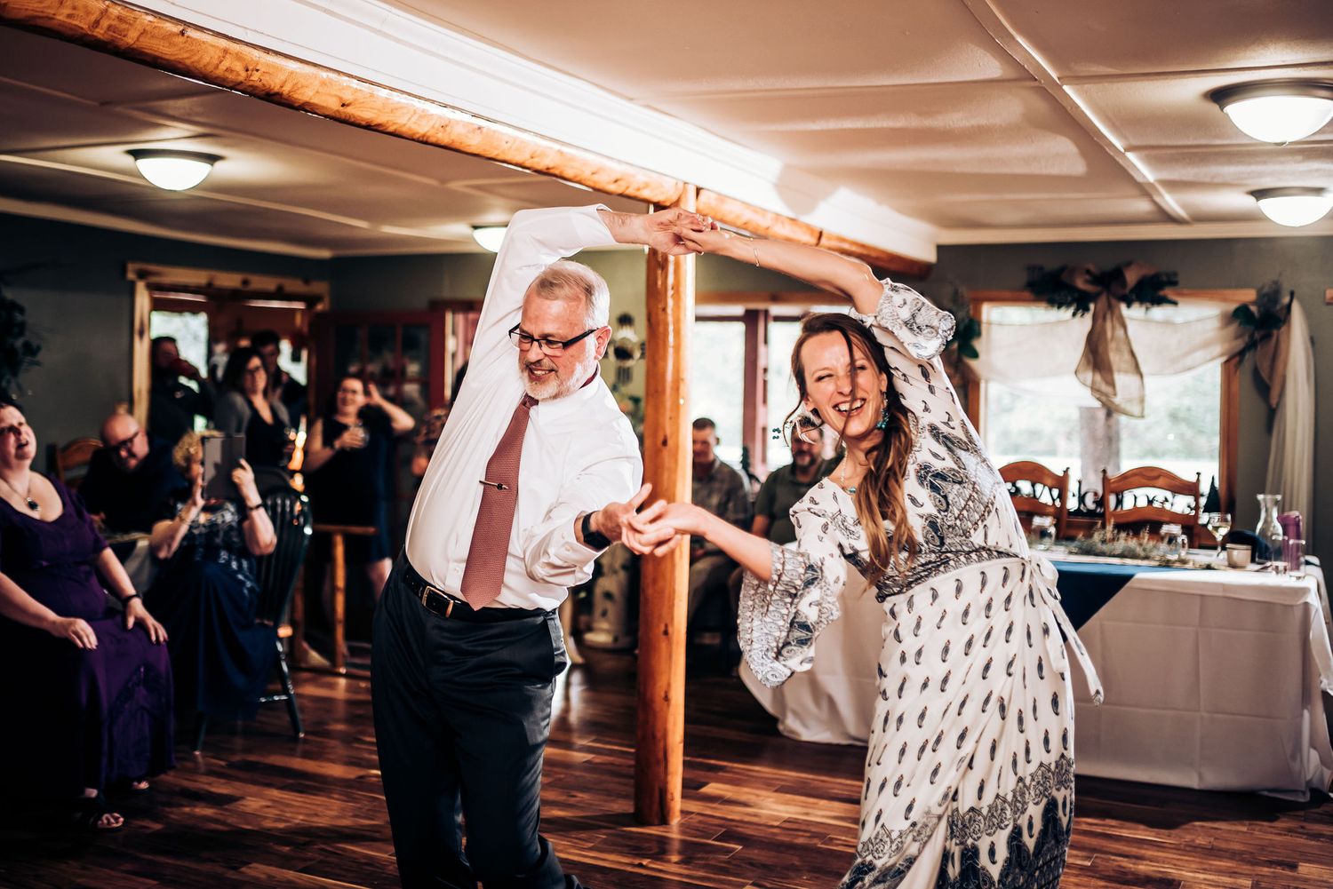 People dancing and celebrating at an indoor reception with exposed wooden beams.