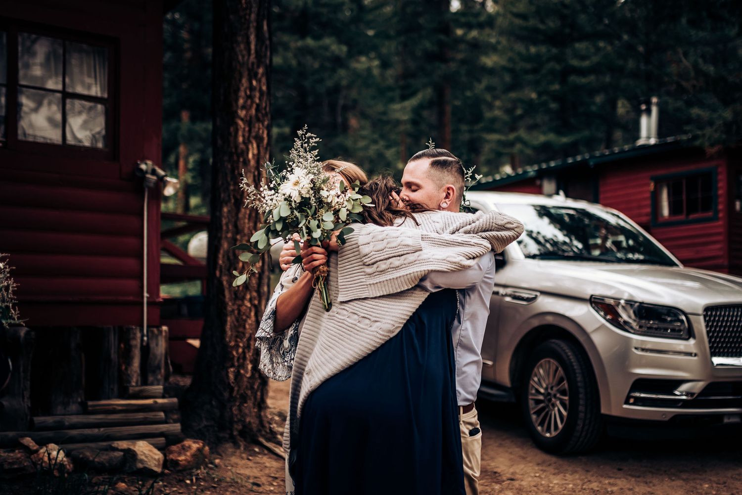 A celebratory embrace in front of a red cabin and car after a forest wedding ceremony.