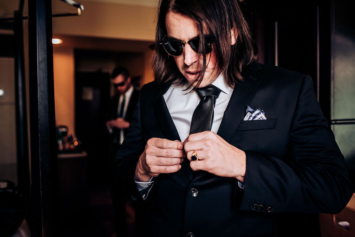 Person in black suit and sunglasses adjusting tie indoors.