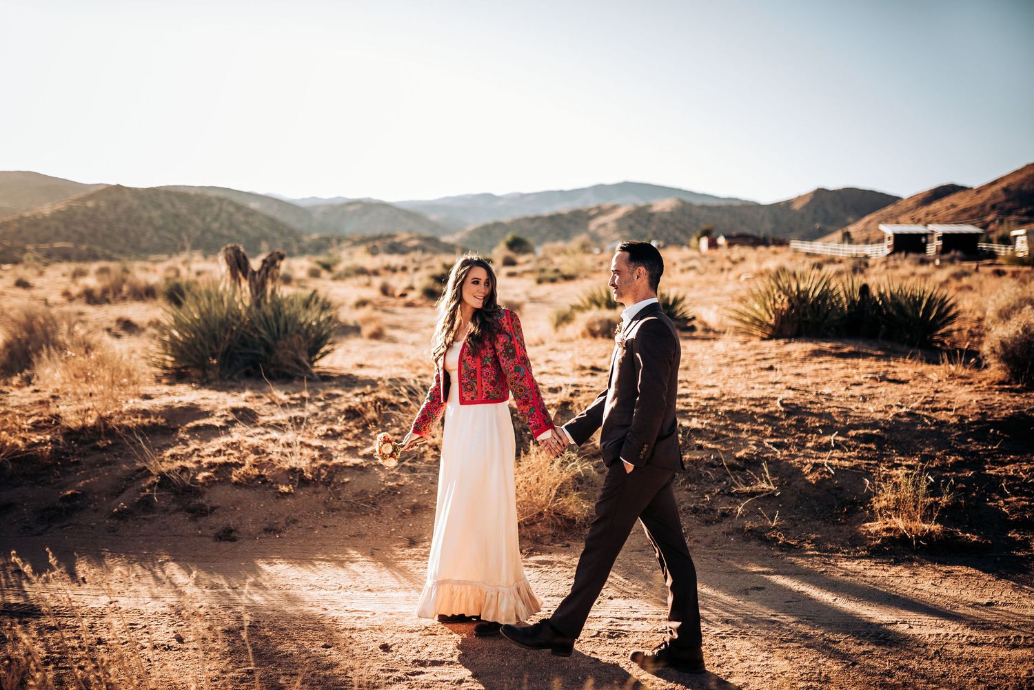 A couple walks hand in hand through a desert landscape with mountains in the background.