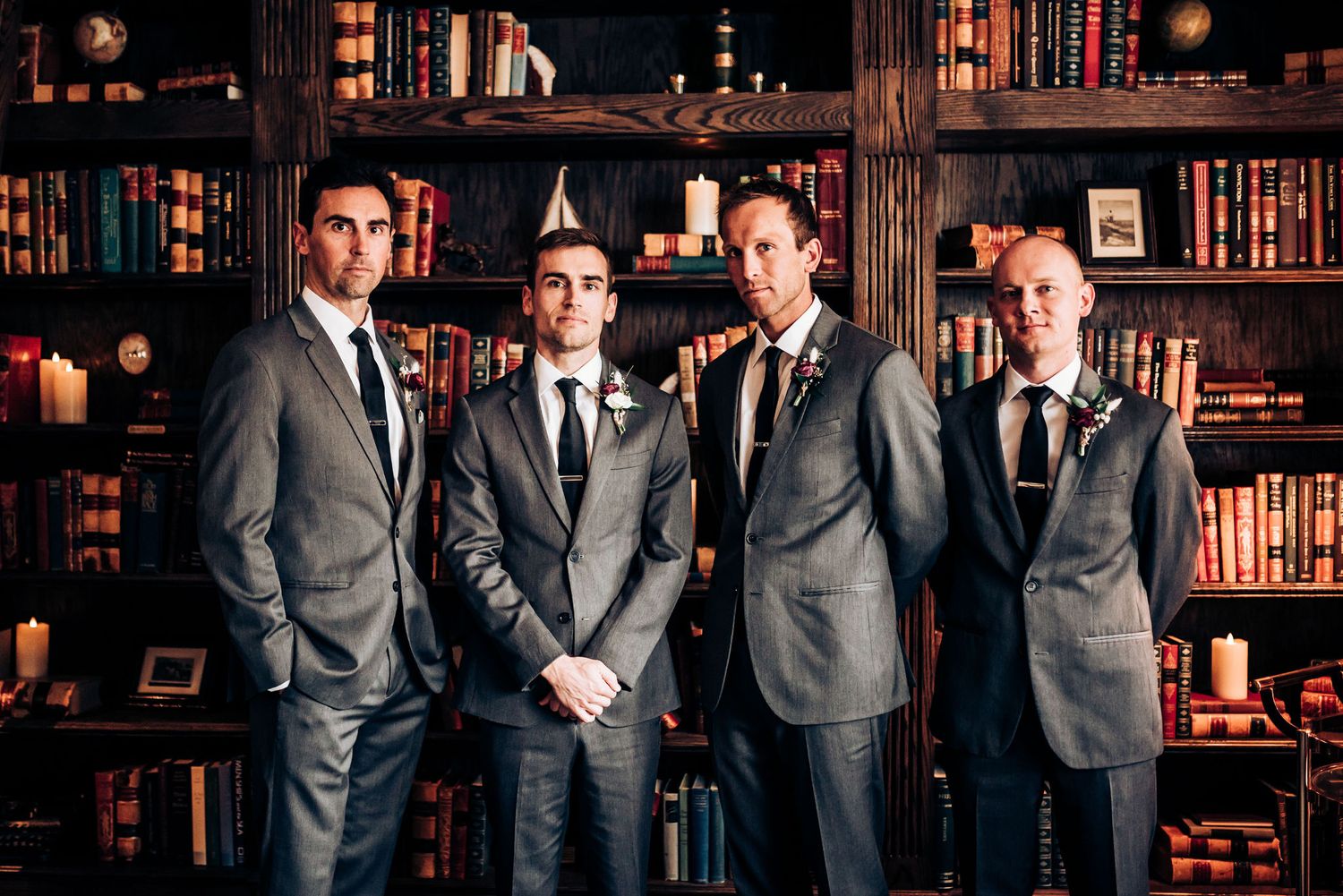 Four groomsmen in matching grey suits stand against dark wood library shelving.