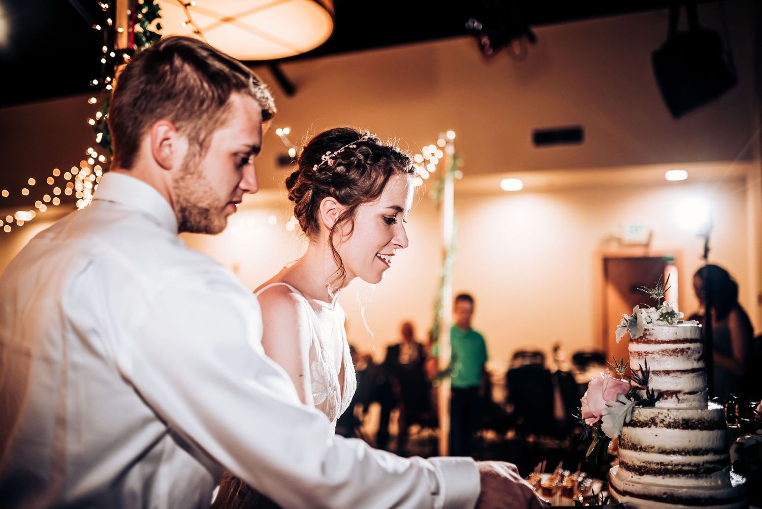 Couple cutting their rustic tiered wedding cake under string lights.