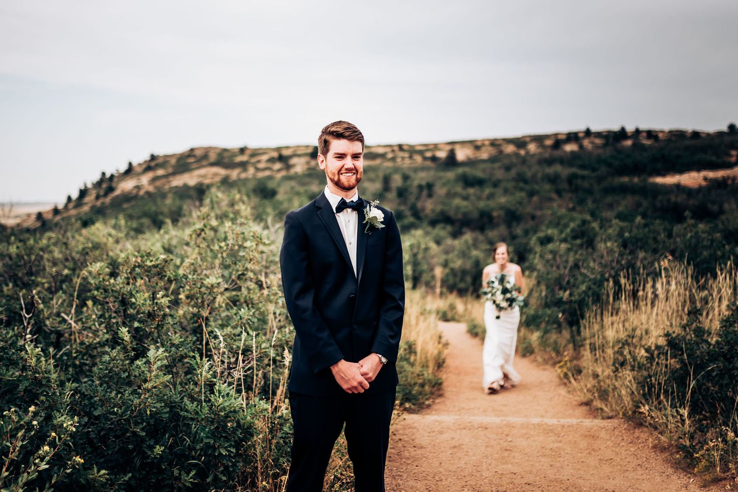 Two figures stand on a scenic mountain trail during a wedding ceremony against a hillside backdrop.