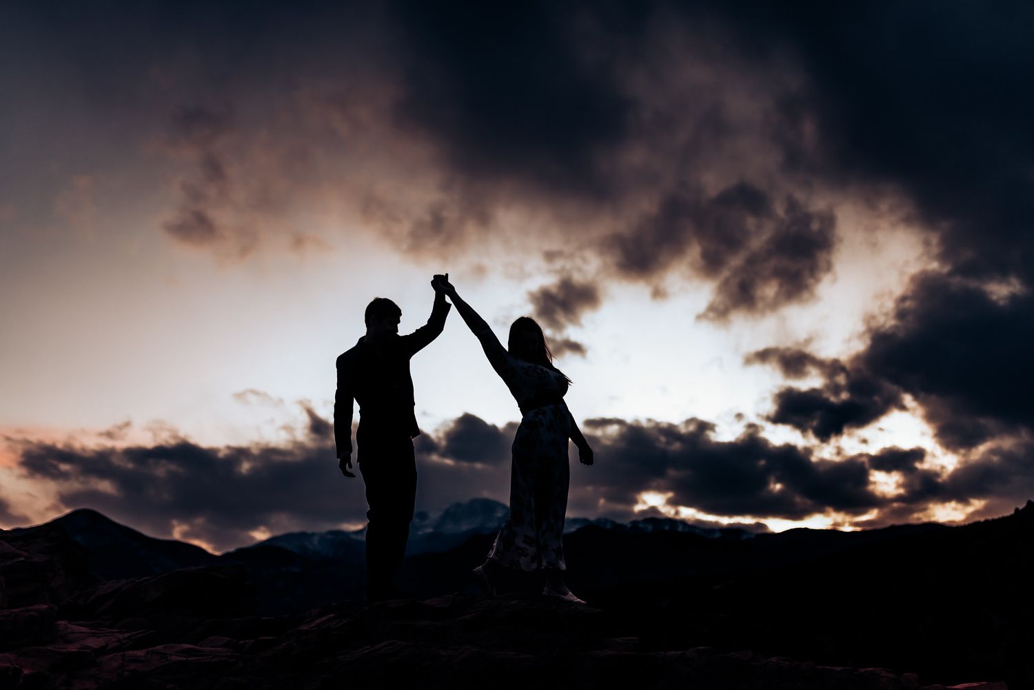 Silhouetted couple high-fives against dramatic sunset sky on mountain peak.