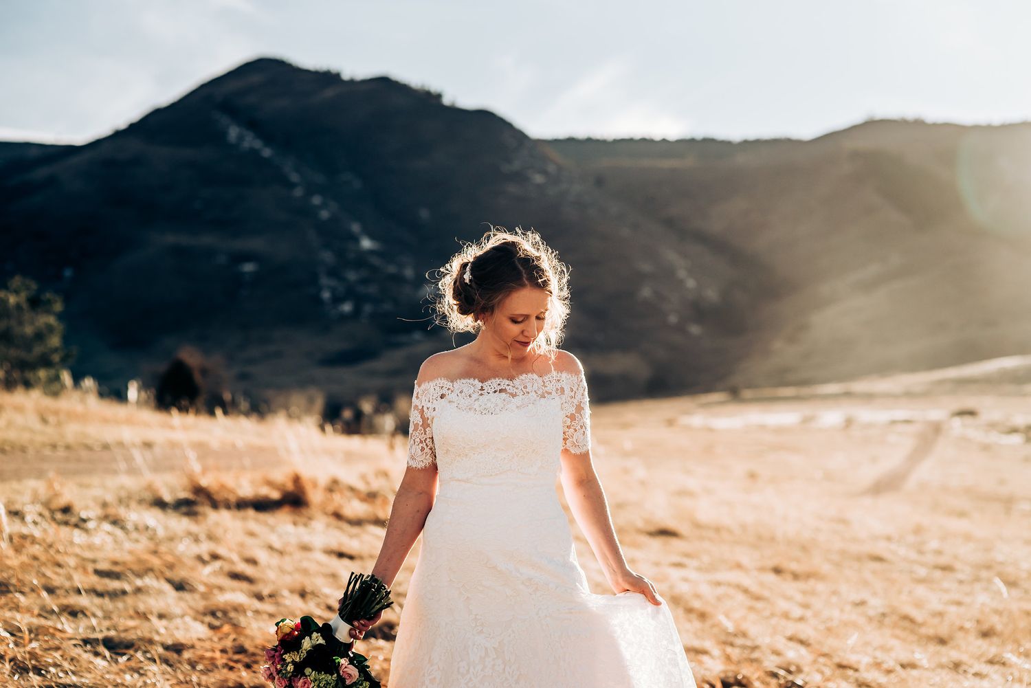 Bride in lace dress walks alone on mountain trail during golden hour.