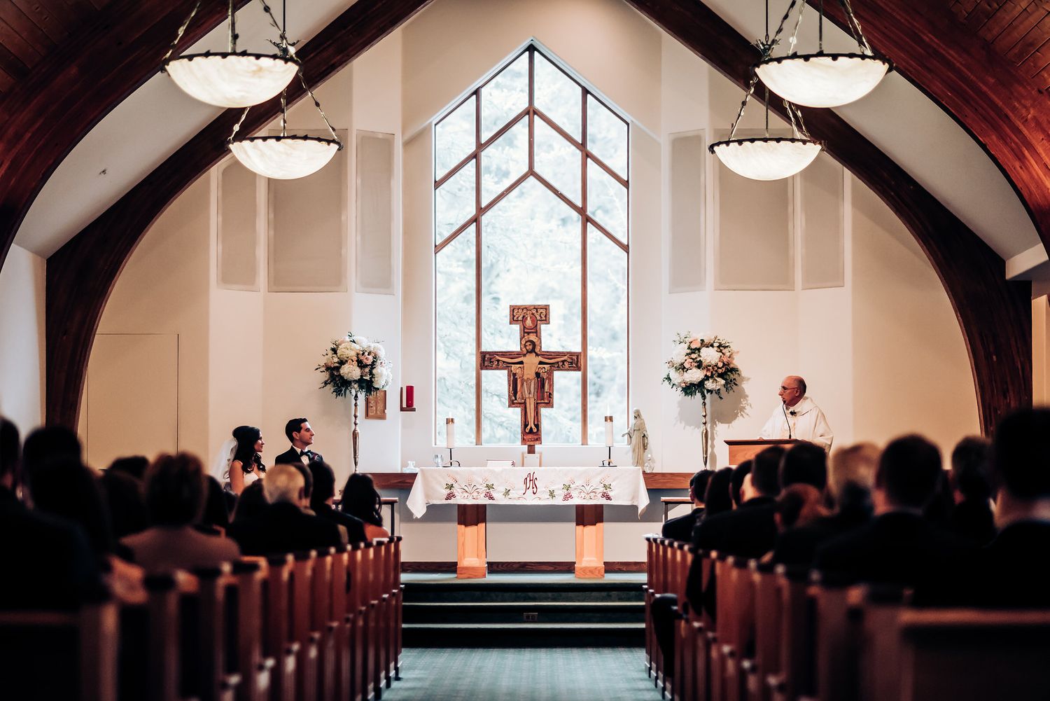 Interior of a church with wooden beams, pendant lights and a large cross at the altar.