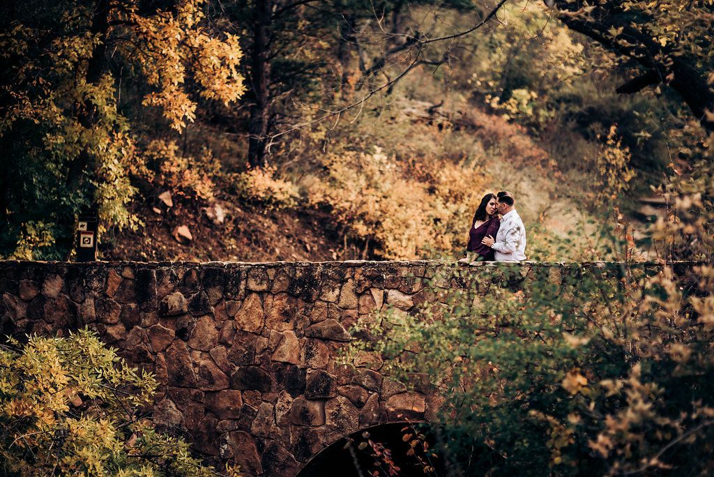Aerial view of couple sitting on rustic bridge surrounded by autumn foliage.