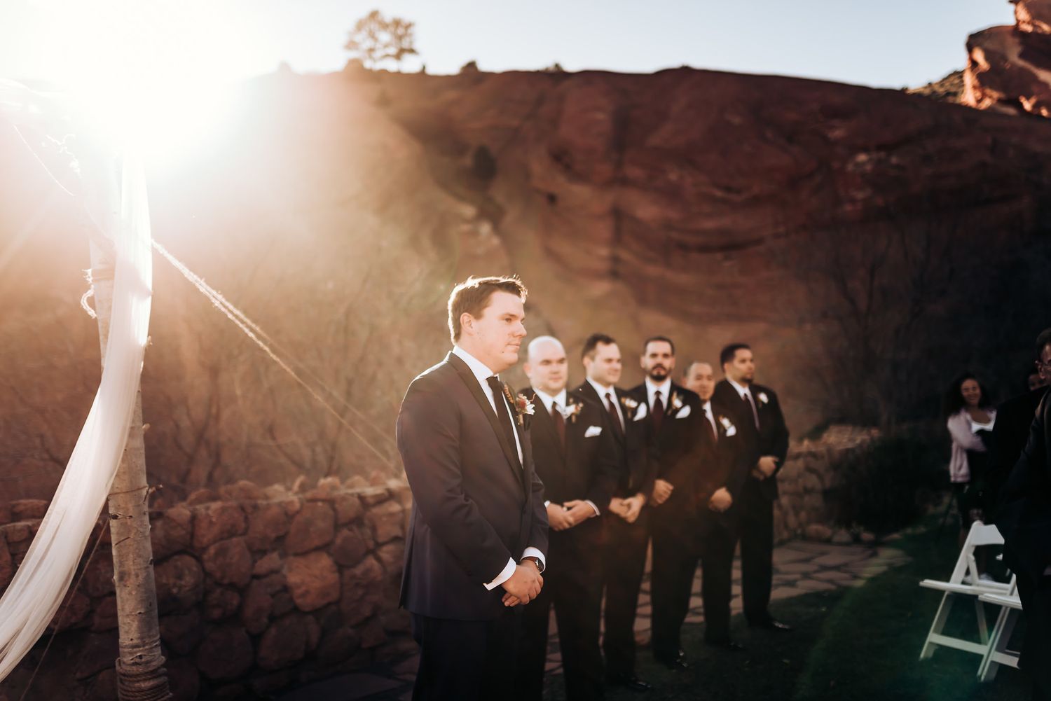 Groomsmen line up at sunset during outdoor wedding ceremony against red rock backdrop.