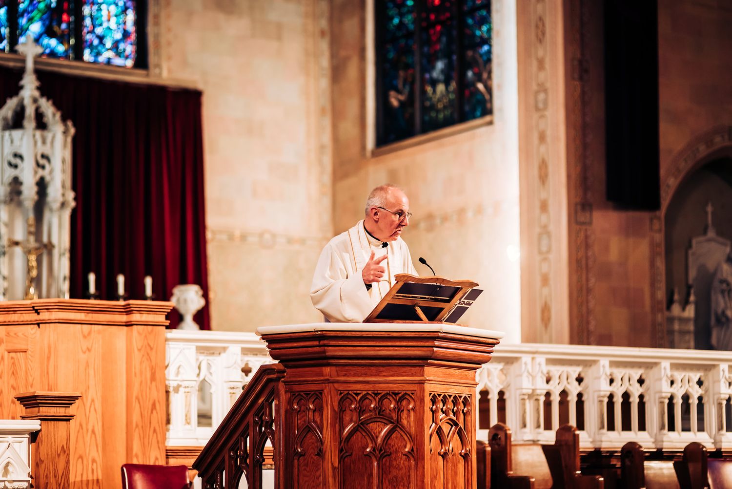 Church minister stands at wooden pulpit delivering sermon with stained glass windows in background.