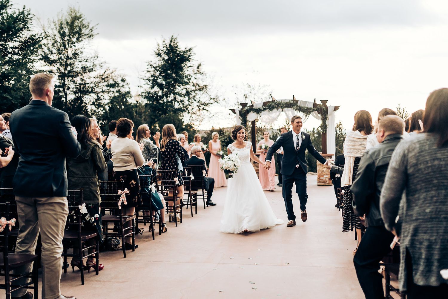 Newlyweds walking down aisle between rows of guests under decorated outdoor pergola.