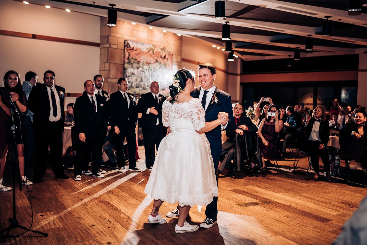 A couple performs their wedding dance on a wooden floor while party guests watch from the sides.