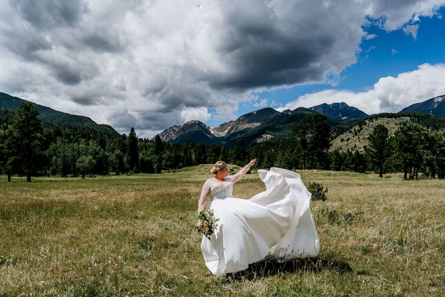 Bride in flowing white dress stands in mountain meadow with dramatic storm clouds overhead.