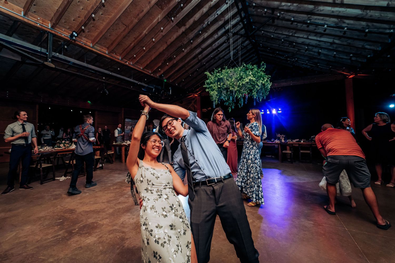 Wedding guests dance enthusiastically on the dance floor under string lights and greenery decor.