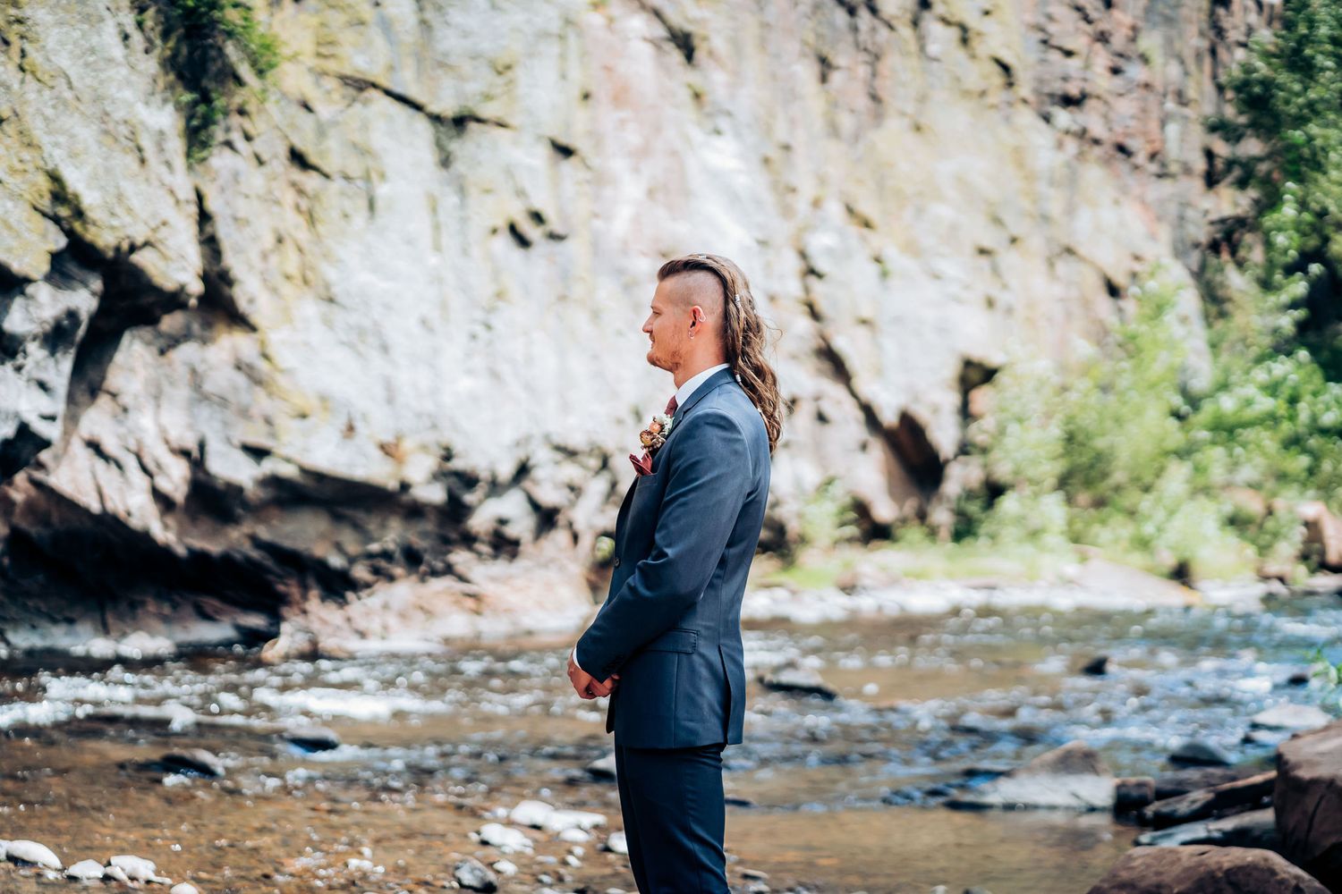 Person in navy suit standing by a rocky riverbank during a wedding ceremony.