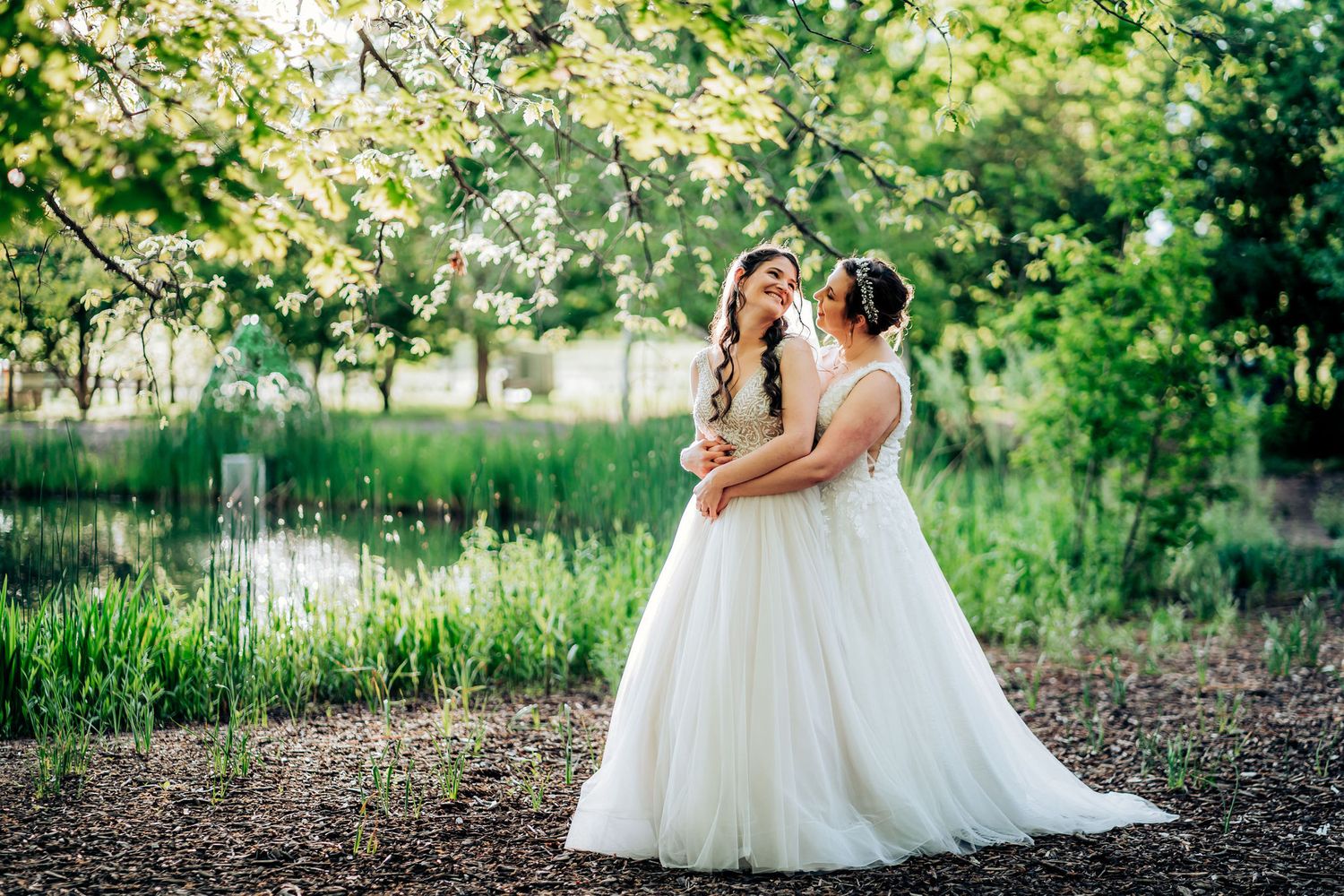 Two brides in flowing white gowns embrace in a serene garden setting during golden hour.