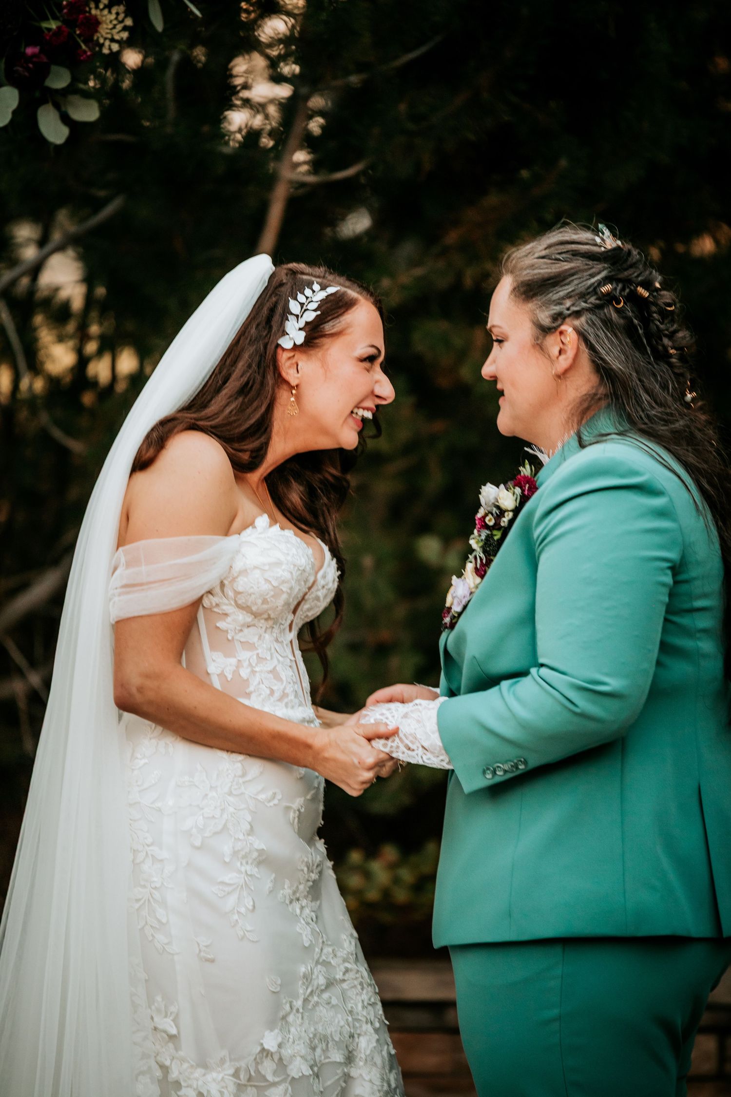 Two brides smile and hold hands facing each other at outdoor wedding ceremony.