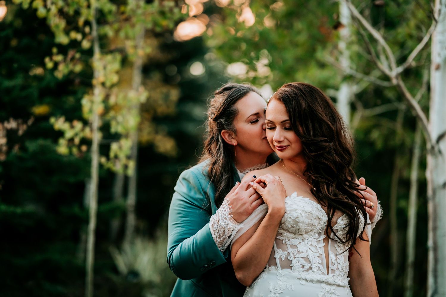 Two brides share a tender kiss outdoors, one in a white lace gown and the other in a teal suit.
