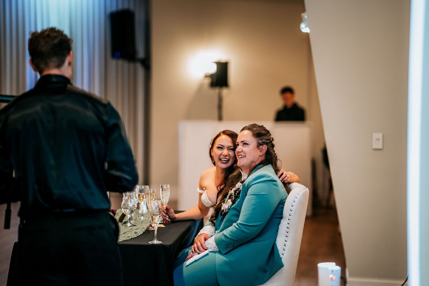 Two brides sit together at decorated head table smiling during wedding reception speeches and toasts.
