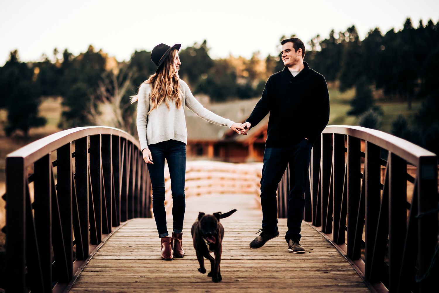 Couple walking with black dog across wooden bridge during sunset.