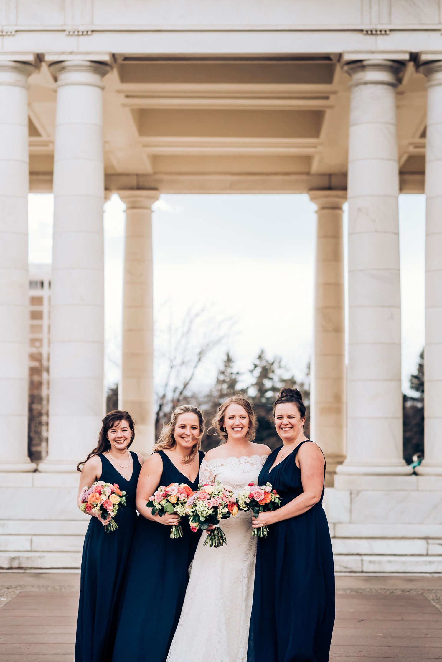 A bridal party with bridesmaids in navy blue dresses holding colorful bouquets under white stone columns.