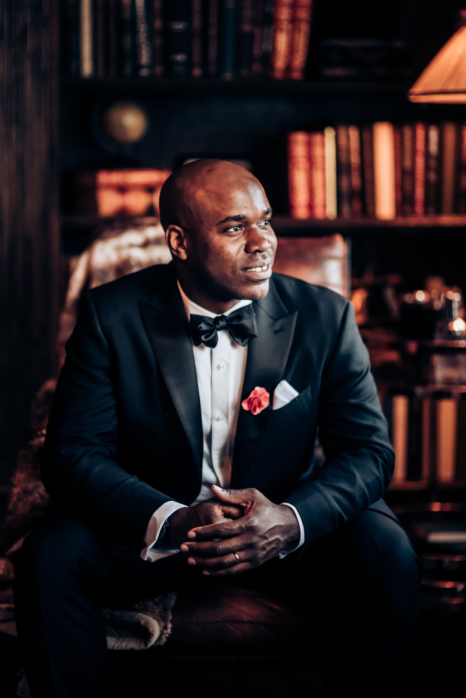 Groom in classic black tuxedo sitting in leather chair against library backdrop.