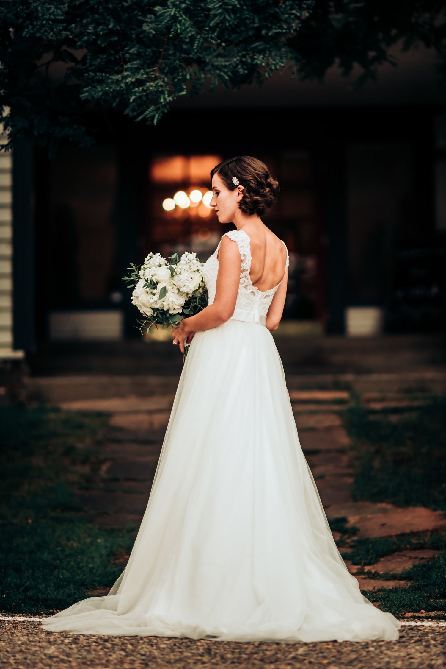 An elegant back view of a wedding gown with lace details and white bouquet against moody lighting.
