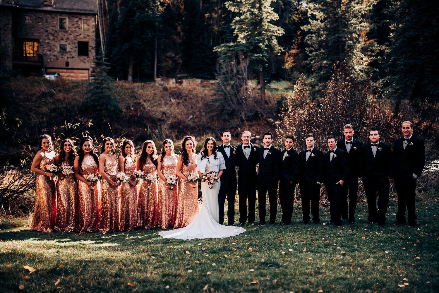 Wedding party in rose gold and black formal attire poses on grass with rustic building backdrop.